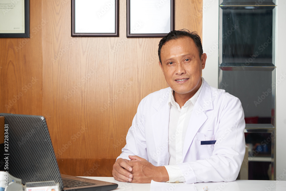 Portrait of smiling confident doctor in coat sitting at his workplace in front of laptop Portrait of smiling confident doctor in coat sitting at his workplace in front of laptop