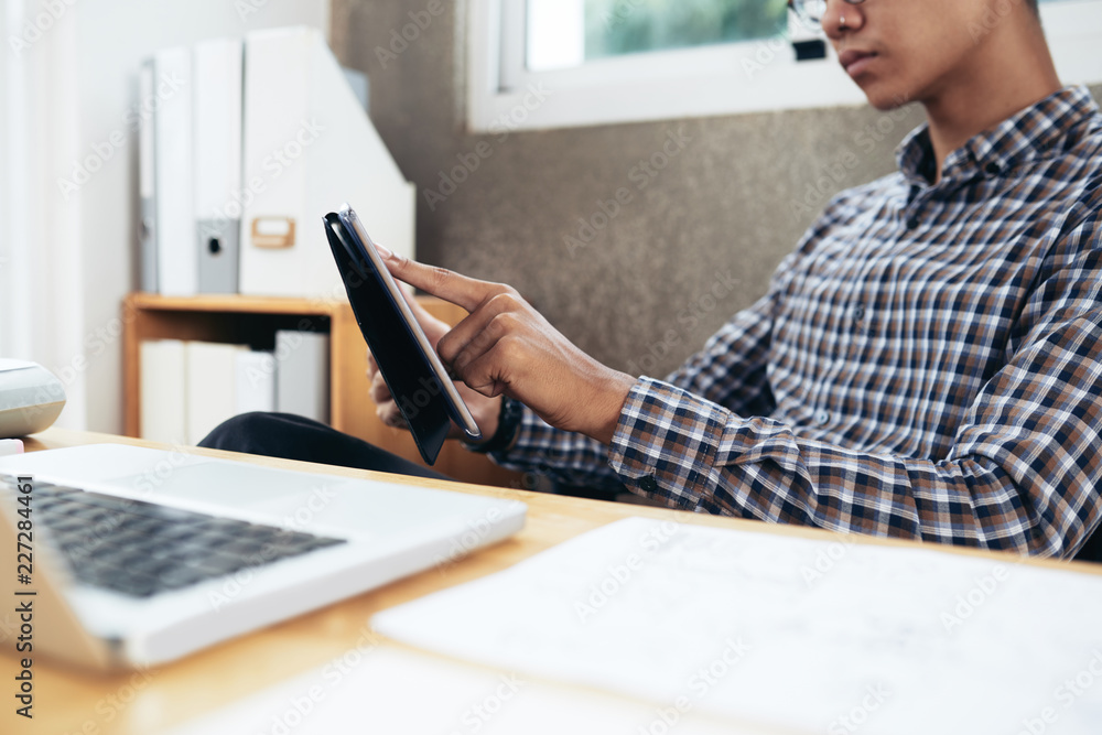 Crop shot of unrecognizable Asian man in checked shirt sitting at office desk and playing on digital tablet while having break Crop shot of unrecognizable Asian man in checked shirt sitting at office desk and playing on digital tablet while having break
