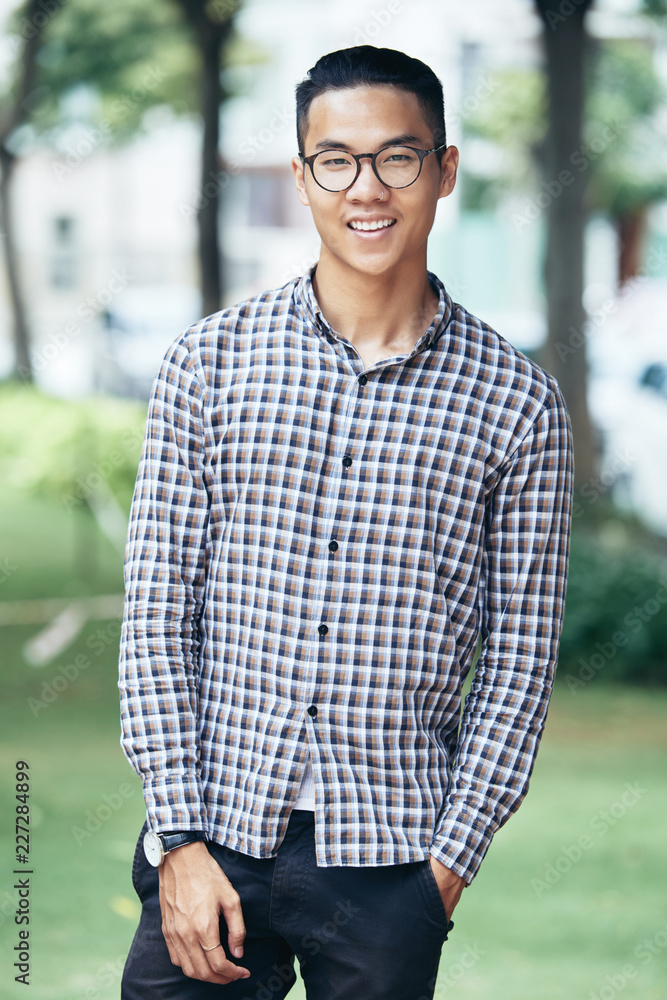 Portrait of young handsome Asian man in checked shirt and round glasses  standing outdoors and smiling at camera happily Stock Photo | Adobe Stock