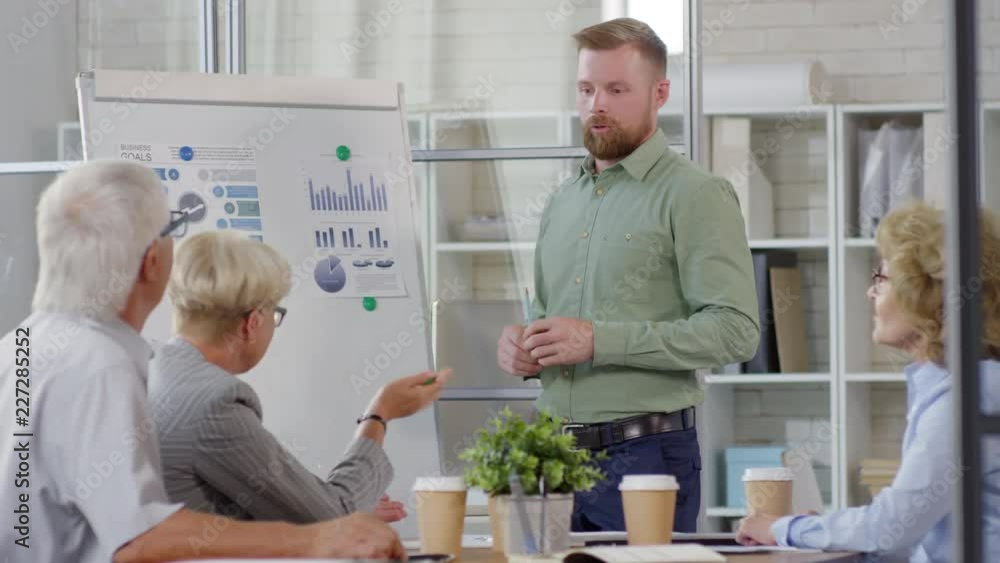Young bearded businessman standing by flipchart in the office and giving presentation about financial growth to team of senior professionals Young bearded businessman standing by flipchart in the office and giving presentation about financial growth to team of senior professionals