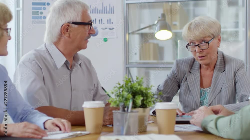Intelligent senior businesswoman sitting at meeting table and explaining something to colleagues while working in the office Intelligent senior businesswoman sitting at meeting table and explaining something to colleagues while working in the office