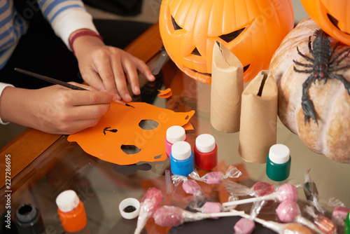 Close-up view of unrecognizable child sitting at table with pumpkins and painting diy paper mask while getting ready for Halloween
