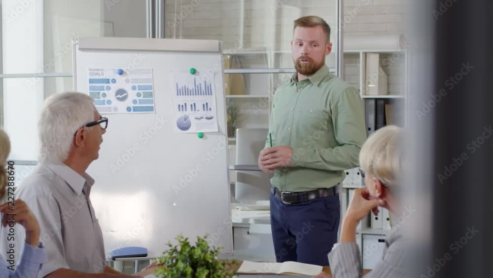 Young professional businessman explaining presentation on flipchart to senior colleagues during office meeting Young professional businessman explaining presentation on flipchart to senior colleagues during office meeting