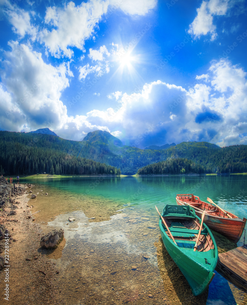 The sun illuminates the light white clouds, shrouded in the morning mist tops of the mountains, the emerald surface of the lake and the still resting boat.