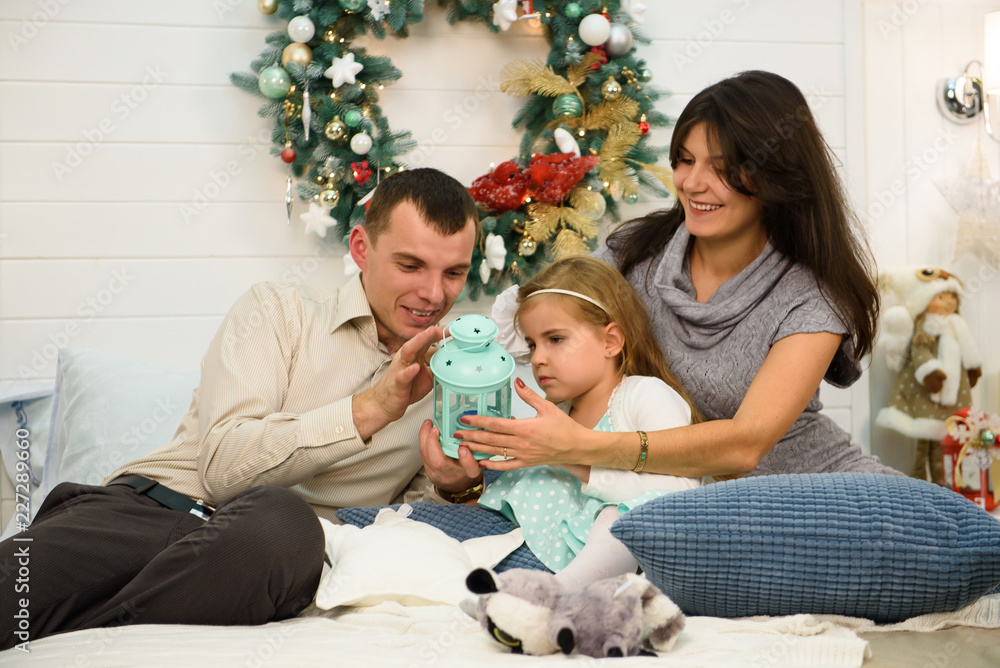 Happy family portrait on Christmas, mother, father and child sitting on bed and lighting a candle at home, chritmas decoration around them Happy family portrait on Christmas, mother, father and child sitting on bed and lighting a candle at home, chritmas decoration around them