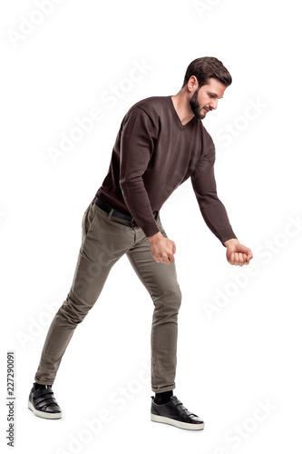 A fit bearded young man in casual clothes tries to pull an invisible rope with all his strength from low grip.