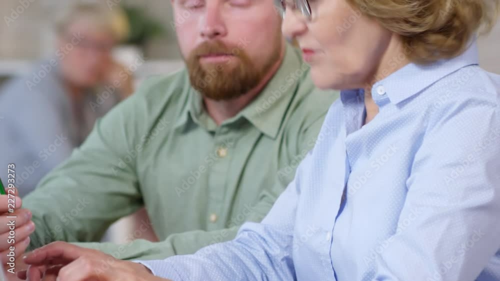 Tilt up of middle aged businesswoman typing on laptop at office desk and speaking with young bearded employee Tilt up of middle aged businesswoman typing on laptop at office desk and speaking with young bearded employee