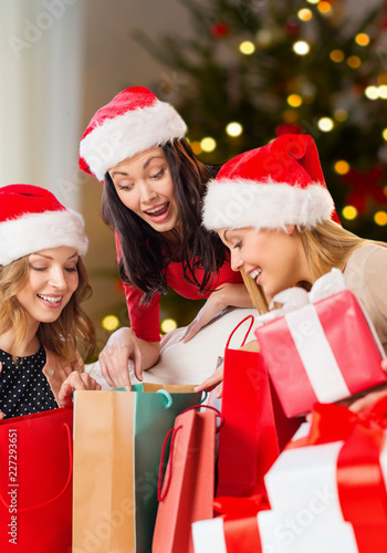 holidays and people concept - women in santa hats with gifts and shopping bags over christmas tree lights background
