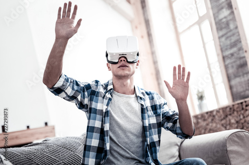 Virtual reality. Calm young man sitting on the sofa and putting his hands up while wearing virtual reality glasses