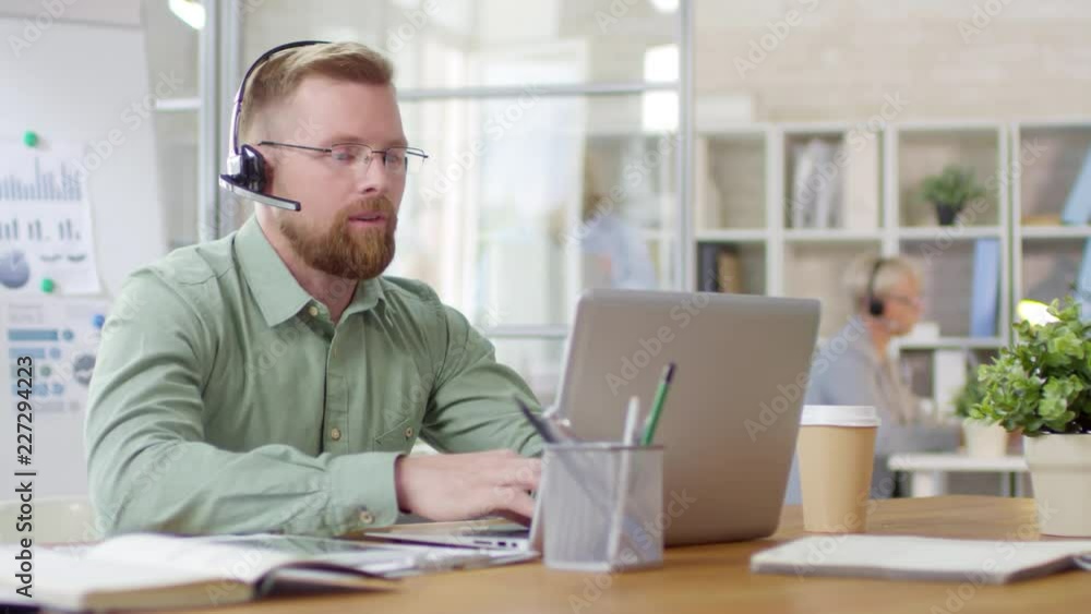 Male call center manager in headset typing on laptop and talking via video call while working at desk in the office Male call center manager in headset typing on laptop and talking via video call while working at desk in the office