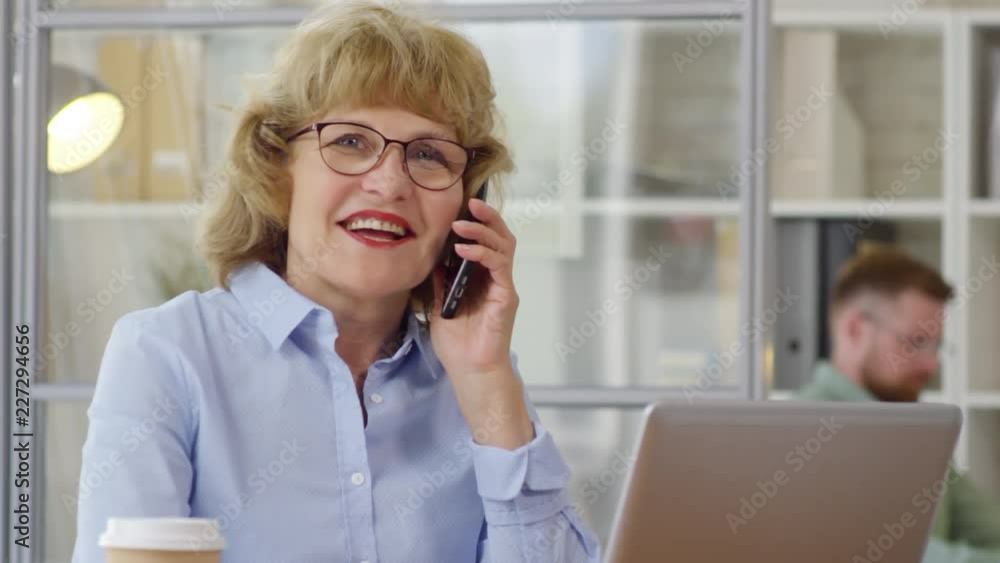 Senior businesswoman in eyeglasses smiling and talking on cell phone while working in the office Senior businesswoman in eyeglasses smiling and talking on cell phone while working in the office