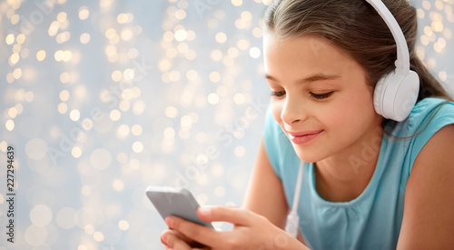 children, technology and people concept - close up of happy girl with smartphone and headphones listening to music over festive lights background