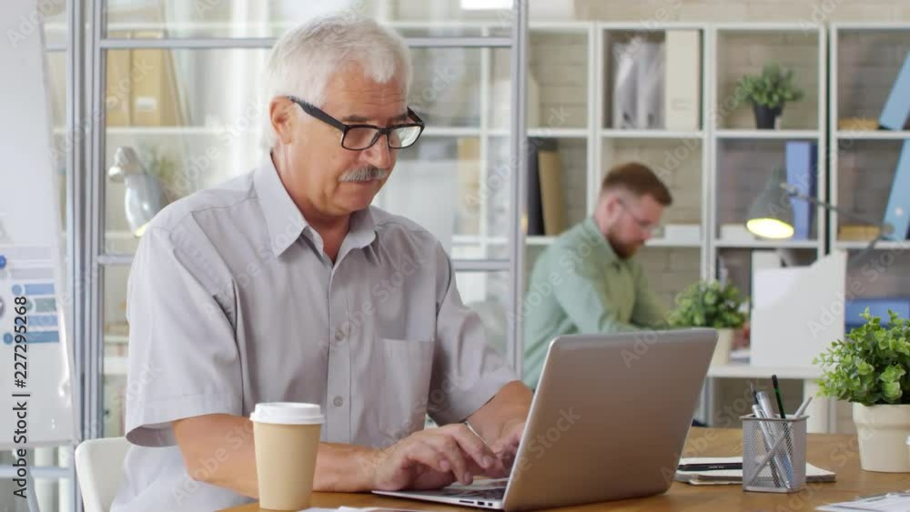 Joyous senior businessman in eyeglasses typing on laptop at desk in the office and smiling at camera Joyous senior businessman in eyeglasses typing on laptop at desk in the office and smiling at camera