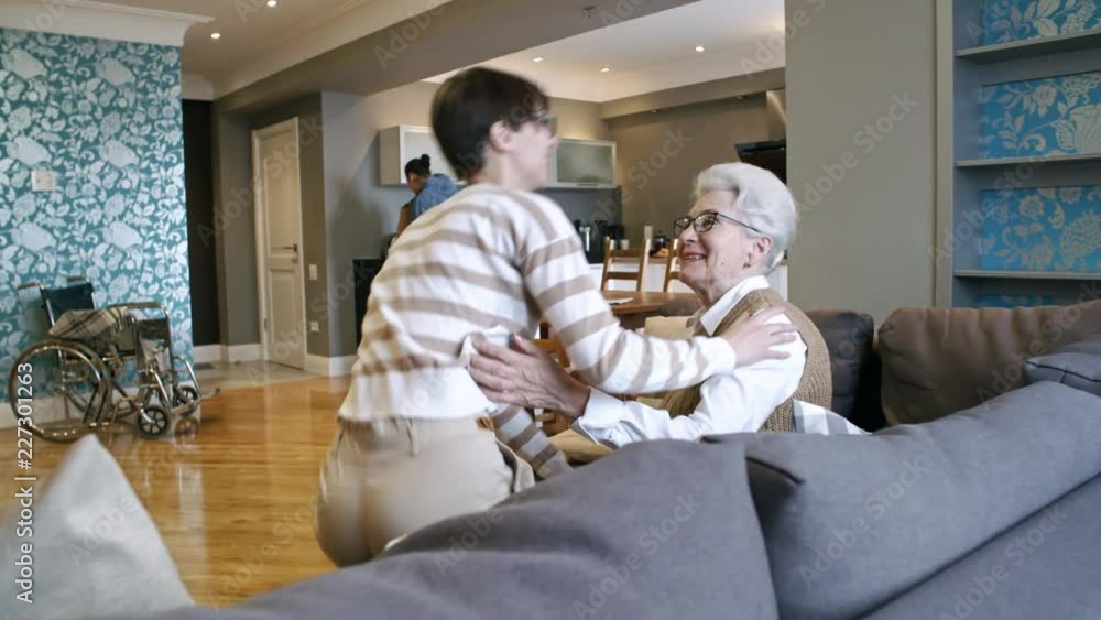 PAN of happy young woman in glasses walking up to smiling elderly lady sitting on sofa and kissing her on cheek, then chatting while man unpacking groceries in background PAN of happy young woman in glasses walking up to smiling elderly lady sitting on sofa and kissing her on cheek, then chatting while man unpacking groceries in background