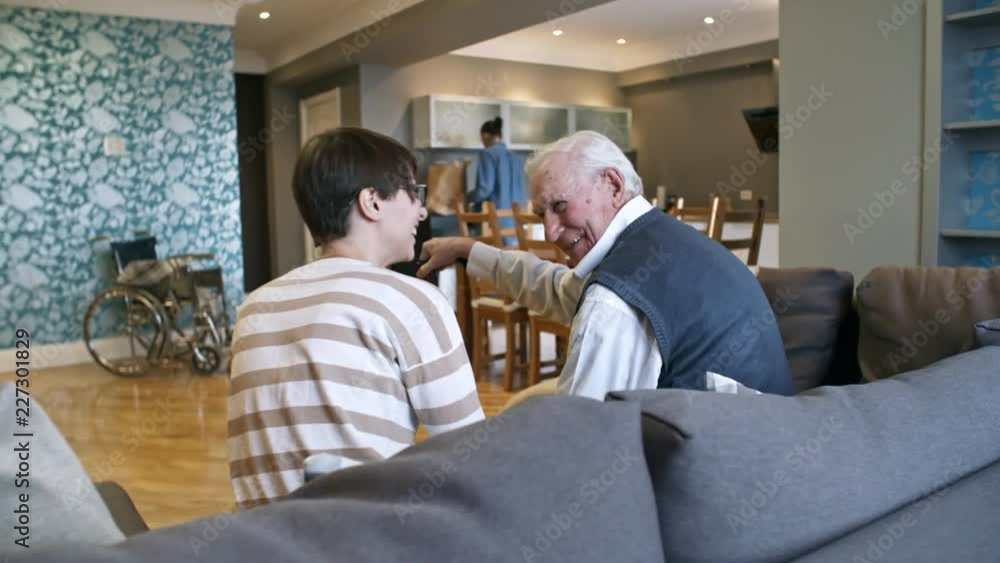 PAN of cheerful female caregiver walking up to cheerful senior man with walking stick sitting on sofa and chatting with him while male volunteer unpacking groceries in kitchen PAN of cheerful female caregiver walking up to cheerful senior man with walking stick sitting on sofa and chatting with him while male volunteer unpacking groceries in kitchen