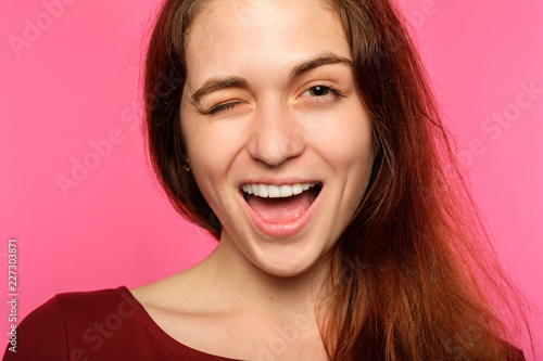 emotion face. smiling cheerful woman pleased with herself. young beautiful brown haired girl winking. portrait on pink background.