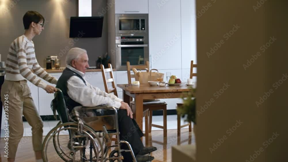 PAN of cheerful young female caregiver in glasses pushing infirm elderly man on wheelchair towards dinner table, then giving him buns with butter for breakfast PAN of cheerful young female caregiver in glasses pushing infirm elderly man on wheelchair towards dinner table, then giving him buns with butter for breakfast