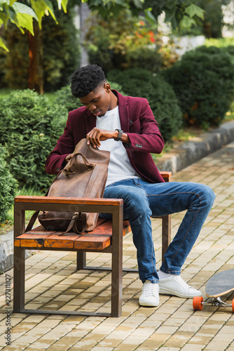 stylish young student with skateboard opening backpack while sitting on bench on street