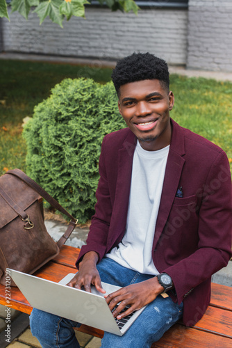 smiling young african american student using laptop on street and looking at camera