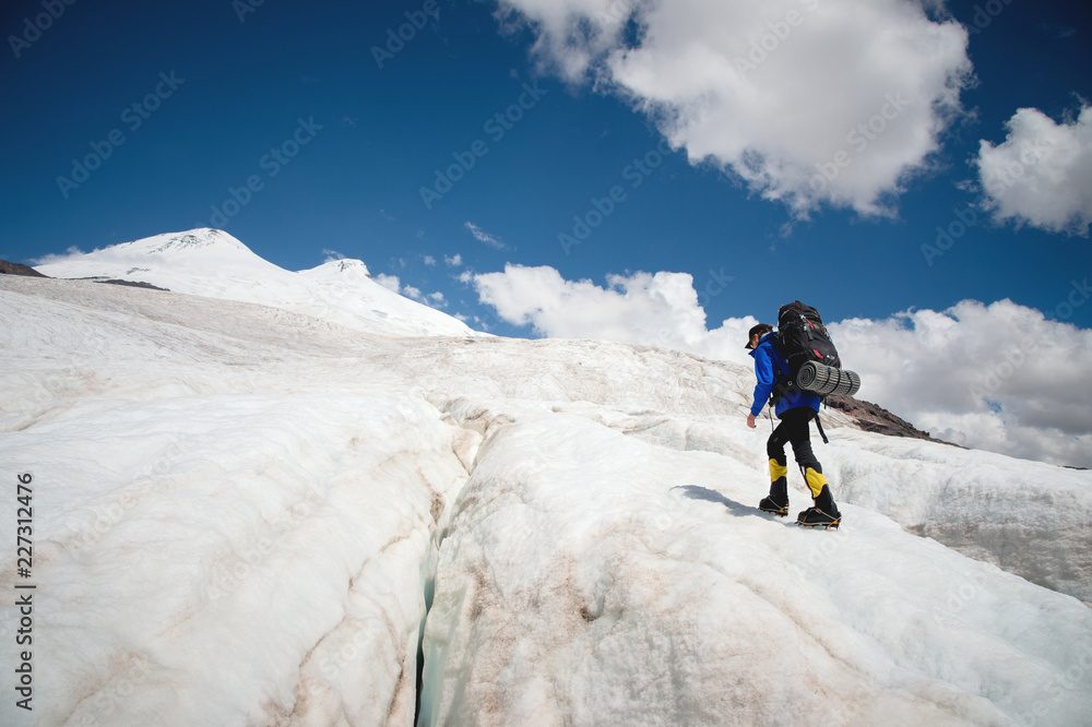 A mountaineer with a backpack walks in crampons walking along a dusty glacier with sidewalks in the hands between cracks in the mountain