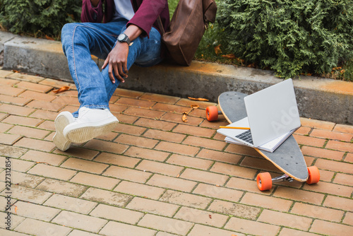 cropped shot of freelancer sitting on curb on street near laptop on skateboard