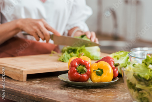 cropped image of woman preparing salad for dinner and cutting cabbage at home, bell peppers on foreground