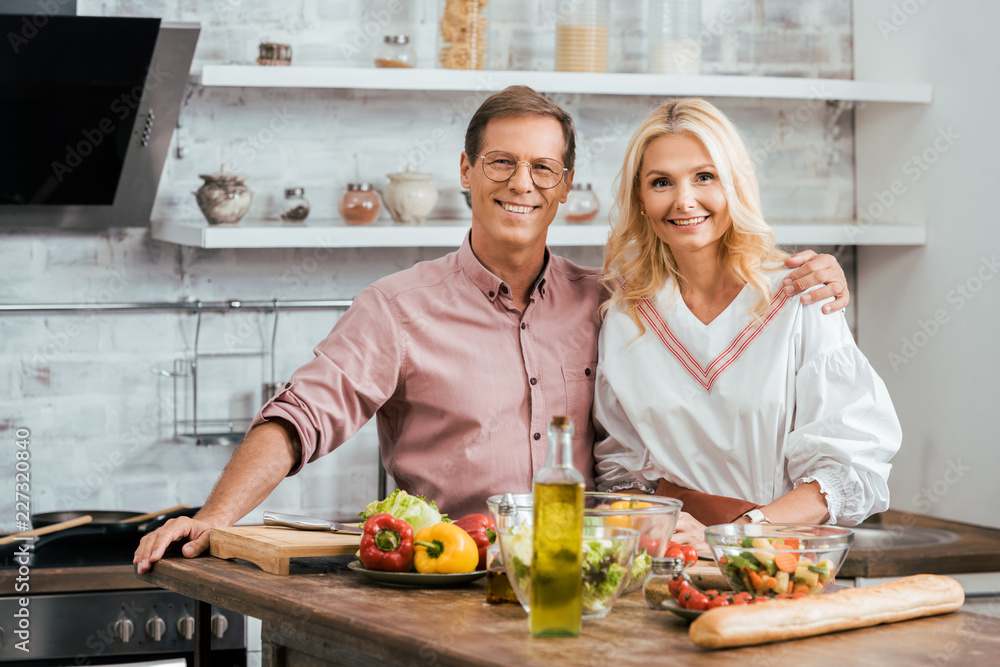 adult couple hugging and looking at camera during salad preparation for dinner at home adult couple hugging and looking at camera during salad preparation for dinner at home