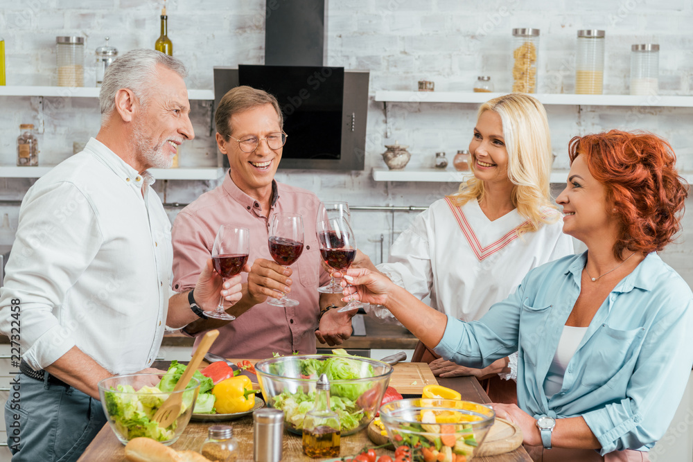 smiling old friends clinking with wineglasses in kitchen smiling old friends clinking with wineglasses in kitchen