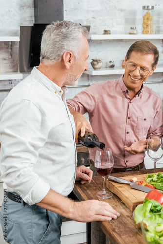 handsome happy man pouring wine to old friend during dinner in kitchen