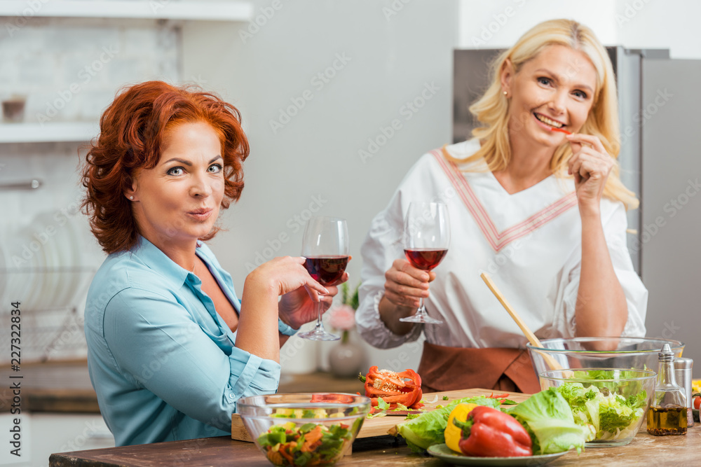 smiling attractive women preparing salad for dinner, holding wineglasses and looking at camera at home smiling attractive women preparing salad for dinner, holding wineglasses and looking at camera at home