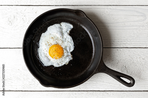 Single fried egg in cast iron frying pan sprinkled with ground black pepper. Isolated on white painted wood from above.