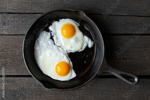 Two fried eggs in cast iron frying pan isolated on dark painted wood from above.