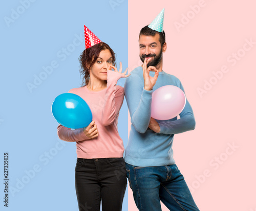 couple with balloons and birthday hats showing an ok sign with fingers on pink and blue background