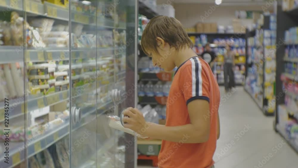 Teenager in supermarket. Caucasian teen boy in red t-shirt choosing fresh food in plastic box from the fridge on store shelves background Teenager in supermarket. Caucasian teen boy in red t-shirt choosing fresh food in plastic box from the fridge on store shelves background