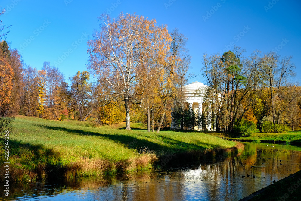 Beautiful autumn sunny landscape in Pavlovsk park with the Slavyanka river, red and orange leaves and the Temple of Friendship behind the trees, Pavlovsk, St. Petersburg. Beautiful autumn sunny landscape in Pavlovsk park with the Slavyanka river, red and orange leaves and the Temple of Friendship behind the trees, Pavlovsk, St. Petersburg.