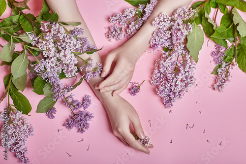 Fototapeta Naklejka Na Ścianę i Meble -  Fashion art hands natural cosmetics women, bright purple lilac flowers in hand with bright contrast makeup, hand care. Creative beauty photo of a girl sitting at table on contrasting pink background