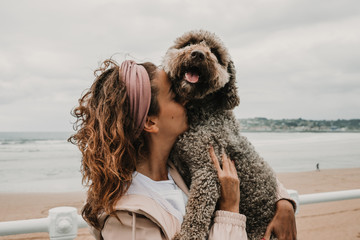 .Young woman wearing a pink coat, playing with her adorable brown spanish water dog on the seafront of Gijon in the north of Spain on a cloudy summer day. Having fun, relaxed afternoon. Lifestyle