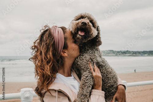 .Young woman wearing a pink coat, playing with her adorable brown spanish water dog on the seafront of Gijon in the north of Spain on a cloudy summer day. Having fun, relaxed afternoon. Lifestyle