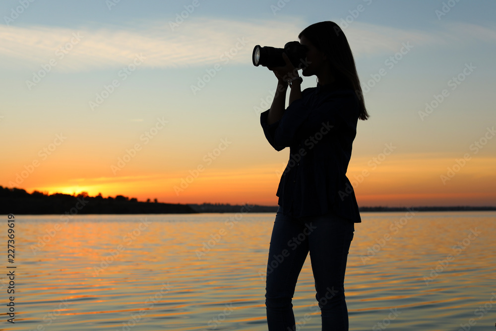 Young female photographer taking photo of riverside sunset with professional camera outdoors