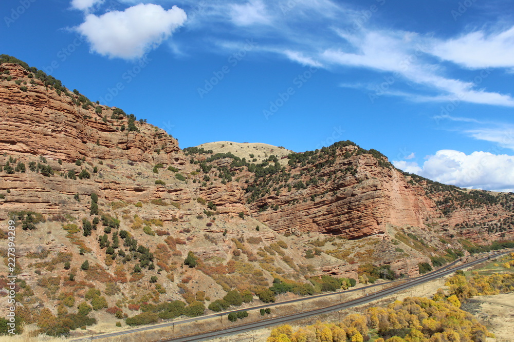 Beautiful hillside with rock faces and shrubs and trees.bright blue sky with a few clouds. a small country road and train tracks run in the foreground