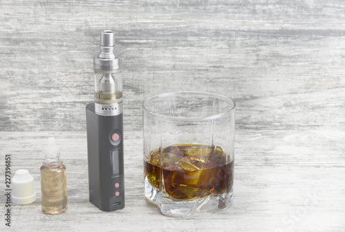 Whiskey in a glass with an e-cigarette and liquid for it on a gray stone background  