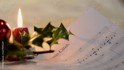 A close well focused shot of a white sheet contaning musical notations for christmas carol kept beside a chistmas holly plant and a burning small candle photo