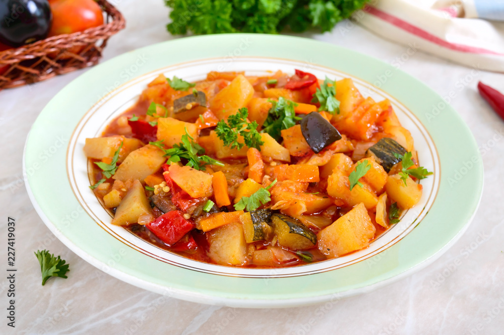 Delicious vegetable stew in a bowl on the table. Stewed potatoes, eggplants, zucchini, onions, carrots in spicy tomato sauce with greens. Delicious vegetable stew in a bowl on the table. Stewed potatoes, eggplants, zucchini, onions, carrots in spicy tomato sauce with greens.