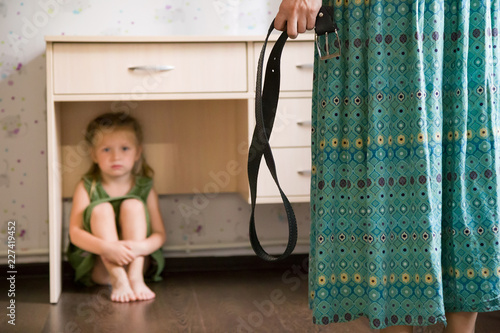 Domestic violence. Mother holding the belt in her hand to punish her small daughter. Scared child hiding under a table and sitting there terrified by physical punishment. Shallow depth of field photo