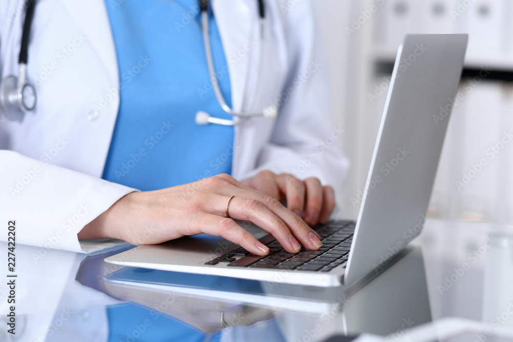 Doctor typing on laptop computer while sitting at the glass desk in hospital office. Physician at work. Medicine and healthcare concept Doctor typing on laptop computer while sitting at the glass desk in hospital office. Physician at work. Medicine and healthcare concept