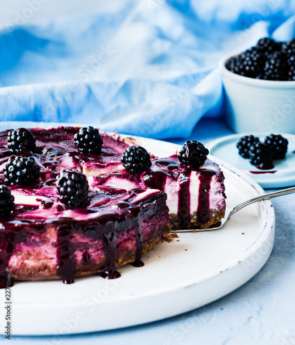 brombeer-cheesecake käsekuchen angeschnitten und fertig zum servieren auf blauem hellen Untergrund auf weißem Teller fertig zum Essen ohne Menschen bei Tageslicht von vorne fotografiert