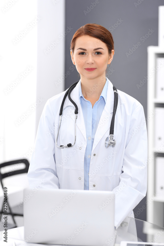 Happy doctor woman at work. Portrait of female physician using laptop computer while standing near reception desk at clinic or emergency hospital. Medicine concept Happy doctor woman at work. Portrait of female physician using laptop computer while standing near reception desk at clinic or emergency hospital. Medicine concept
