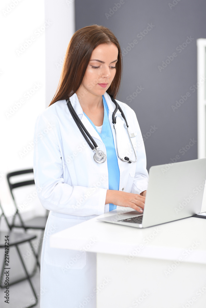 Doctor woman at work. Portrait of female physician using laptop computer while standing near reception desk at clinic or emergency hospital. Medicine and healthcare concept Doctor woman at work. Portrait of female physician using laptop computer while standing near reception desk at clinic or emergency hospital. Medicine and healthcare concept