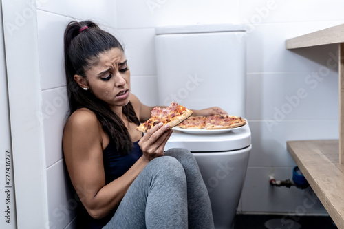 Young beautiful bulimic woman sitting on the bathroom floor eating pizza looking guilty photo