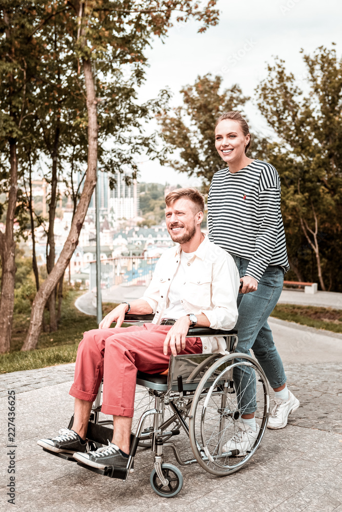 Walk with friend. Positive disabled man sitting in the wheelchair and feeling relaxed while spending time with his reliable girlfriend Walk with friend. Positive disabled man sitting in the wheelchair and feeling relaxed while spending time with his reliable girlfriend